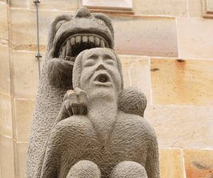 Stone statue on outdoor roof structure of a 12th century French church of a lion-like creature devouring a human with a beard. Both figures have their mouths open. The name of the church is Saint Pierre et Paul, and it's in Rosheim in the region called Bas-Rhin.