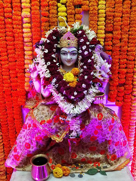 The Hindu goddess Santoshi Mata in a crown, magenta and red robes, with burgundy and white garlands around Her neck, against a background of marigolds.
