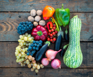 multicolored harvest laid out on an unpainted wooden table: red, green, and magenta peppers, wallnuts, light green, reddish green and dark blue grapes, pink onions, black eggplant, a huge dark and light-green stripeed squash
