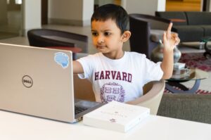 Asking better means knowing that if you don't get what you wanted, you are setting your sights too low. Image of an Indian child with one hand and one finger raised slightly behind to ask a question while looking forward into a laptop in a lounge area. The child is wearing a white Harvard t-shirt with maroon lettering and insignia and has black hair and eyes, looking about six years old. Image credit: Rohit Farmer on Unsplash.